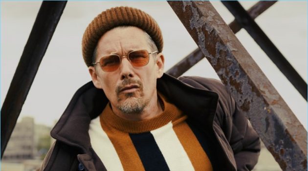 A cool vision, Ethan Hawke wears a BOSS coat, pants, and beanie. Hawke also rocks a Sandro sweater and Garrett Leight sunglasses.