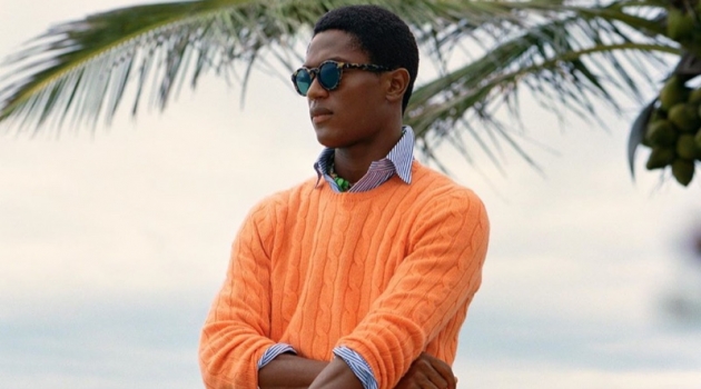 Hamid Onifade sports a bright orange cable-knit sweater with a striped oxford shirt and frayed white shorts by POLO Ralph Lauren.