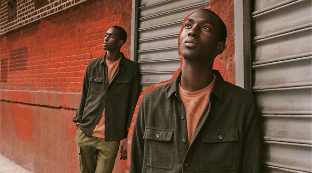 Fall Layers: Embracing autumnal tones, Macky Dabo layers in menswear from East Dane. Posing against a colorful brick wall and metal door, Macky wears a Banks Journal brushed herringbone shirt jacket and waffle fleece sweatshirt. He also models Alex Mill cargo pants with Birkenstock Soft Arizona suede sandals.