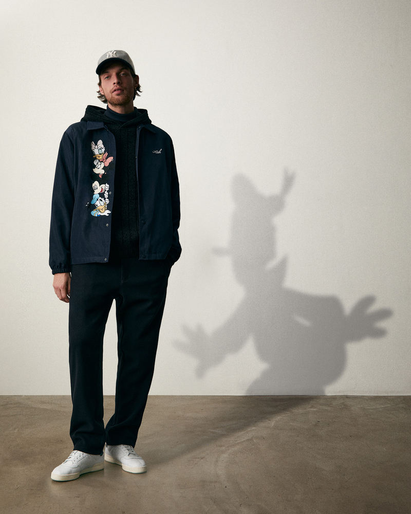 Wearing a look from the Kith for Mickey & Friends collection, model Rocky Harwood stands confidently in a navy jacket adorned with classic Disney characters.