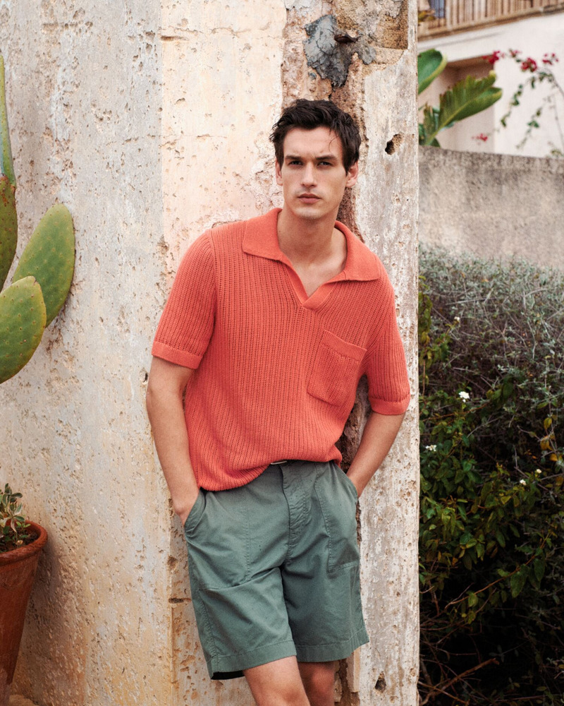 Jegor Venned leans against a rustic wall in GANT's summer 2024 collection, pairing a coral knit polo with sage green shorts.