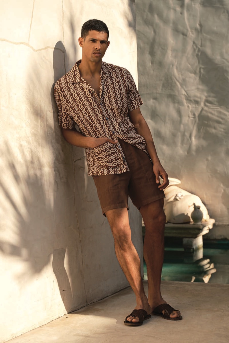 Against a rustic backdrop, Jonas Barros leans effortlessly in a geometric Dobby short-sleeve camp collar shirt paired with 7" Italian linen beach shorts and Tuscan leather woven crisscross sandals.