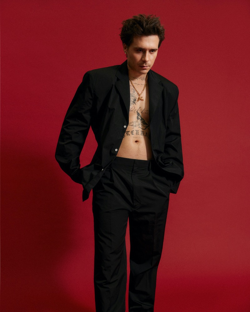 Brooklyn Beckham wears an open Prada blazer, revealing his tattooed torso against a bold red backdrop.