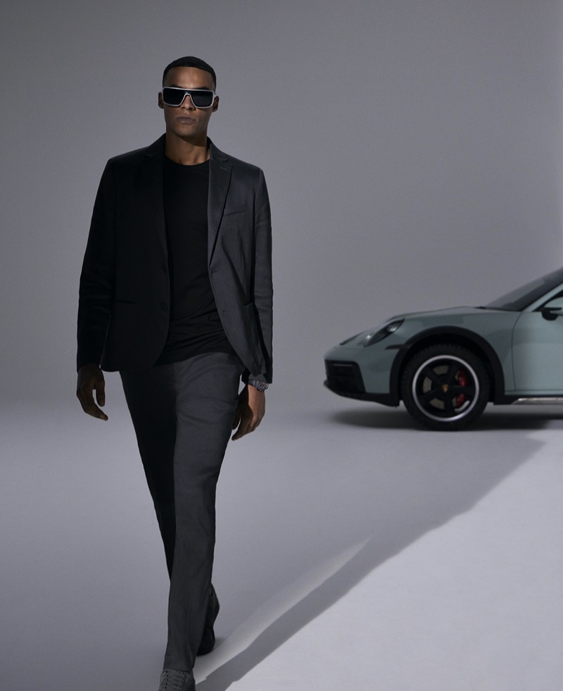 Gaetan Wathelet walks confidently in a dark ensemble, complemented by the striking Porsche Design Iconic Curved P8952 sunglasses and a luxurious Porsche in the backdrop.