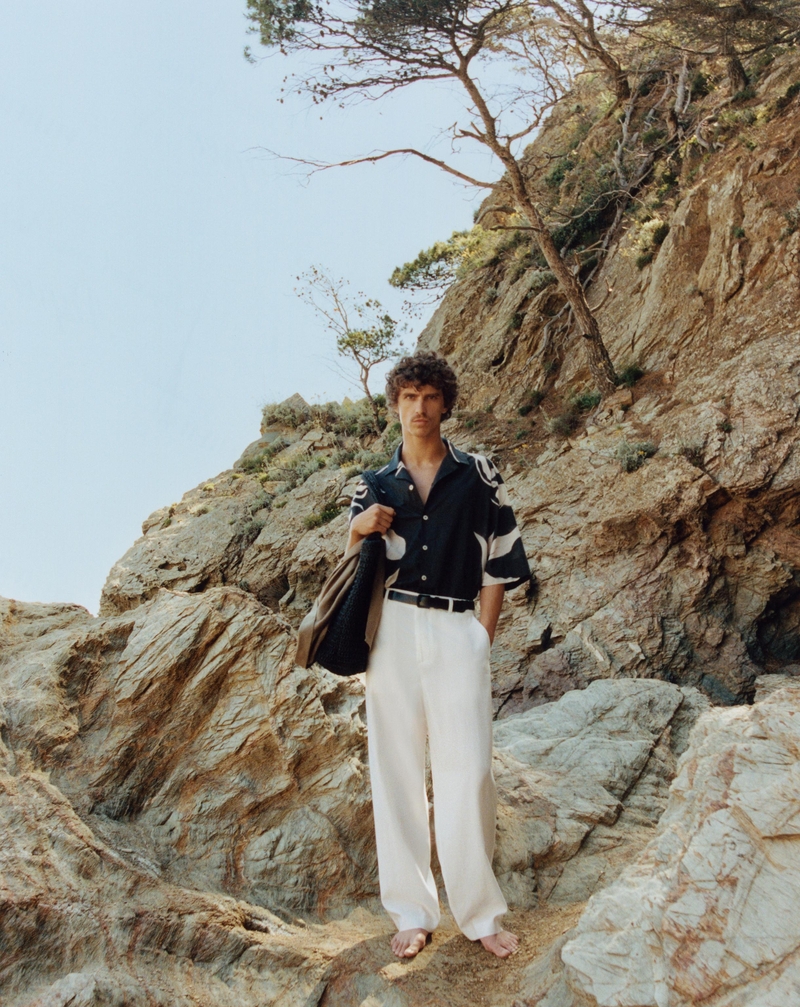 Grzegorz Chanko poses on a rocky terrain, wearing a black floral print shirt with crisp white trousers.