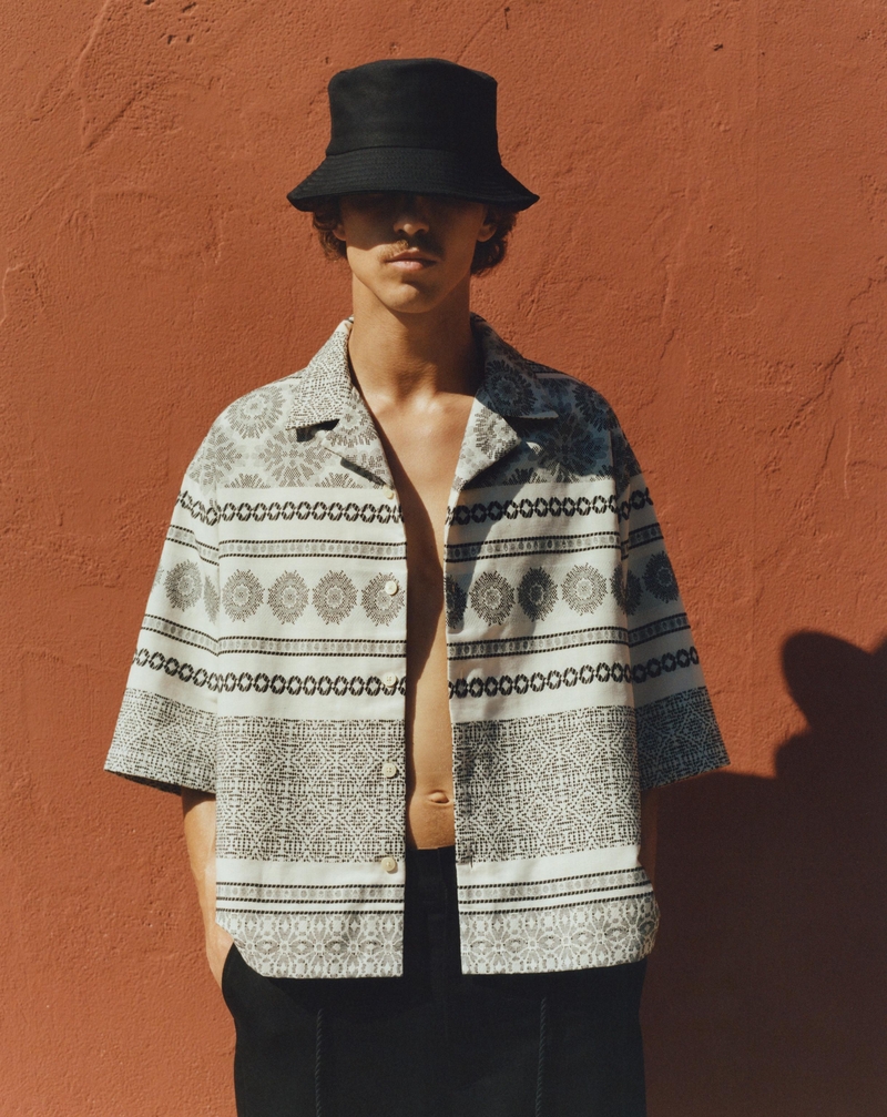 Against a terracotta wall, Grzegorz Chanko sports a patterned short-sleeve shirt and a stylish black bucket hat. 